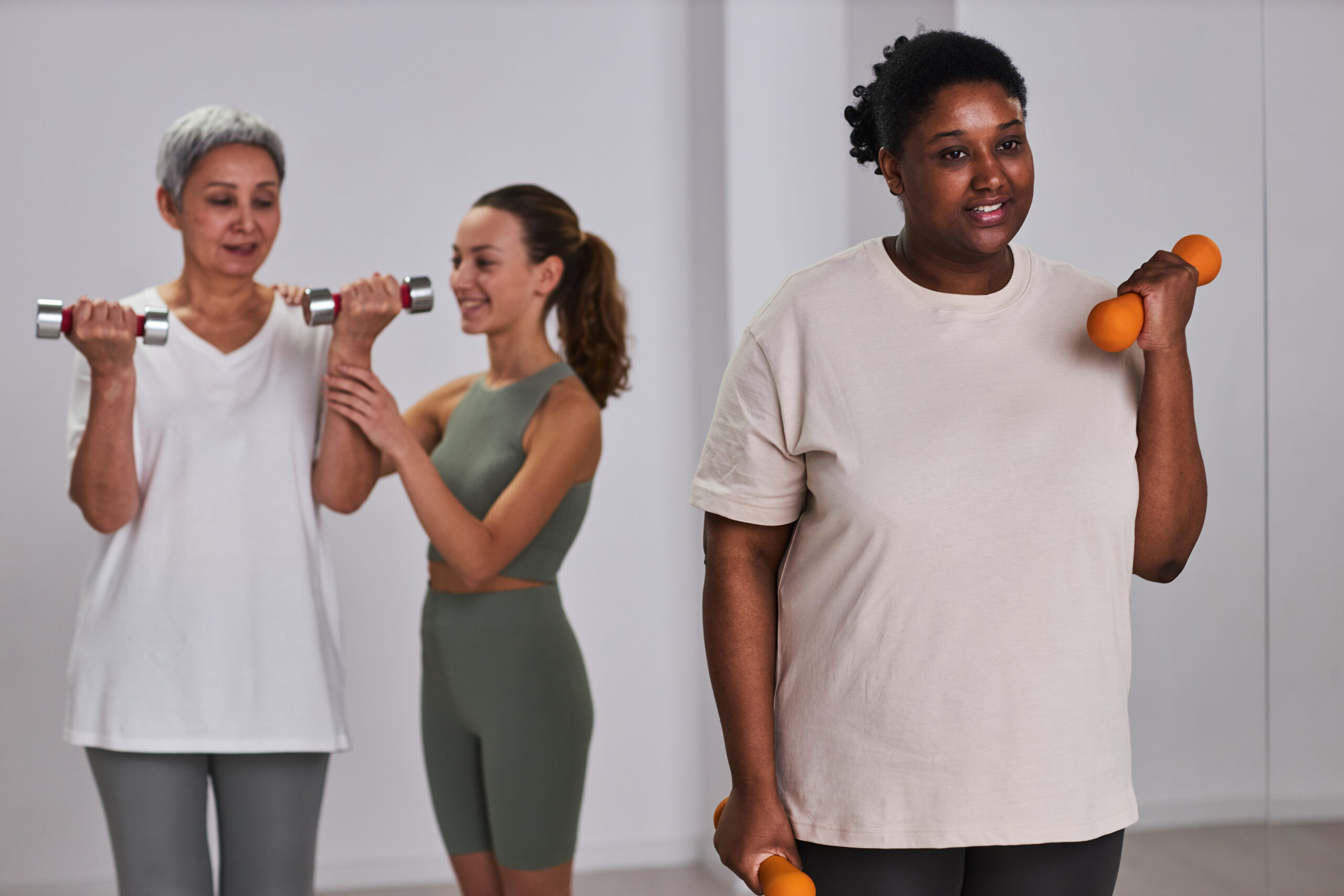 Overweight woman exercising with dumbbells in health club with instructor training mature woman in background