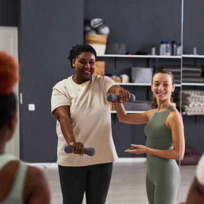 Young instructor showing exercises with dumbbells to overweight woman during training in class